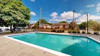 Located at 7820 Lexington Avenue in Cleveland, Ohio, this outdoor recreational area centers around a generously sized swimming pool with clear, blue-green water. The pool deck is finished in smooth concrete and offers ample space for lounging and movement around the perimeter. Depth markers are visible along the pool's edge, indicating a gradual transition from shallow to deeper water, with entry provided via metal handrail steps.

A standout feature of the space is the wooden pergola structure situated to one side of the pool area, providing shaded seating beneath its slatted roof. Colorful picnic-style tables and benches are positioned throughout the deck, offering comfortable gathering spots for visitors. Mature trees, including a notably large deciduous tree adjacent to the pool, provide natural shade and contribute to a pleasant, park-like atmosphere.

The entire area is enclosed by a black metal fence, ensuring safety and privacy. The surrounding landscape includes maintained shrubbery, ornamental grasses, and green foliage, adding to the inviting outdoor ambiance. A dedicated support building with restroom and utility access is adjacent to the pool. The space opens to views of the broader residential community, with brick buildings visible in the background under open, partially cloudy Midwestern skies.