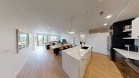 This contemporary single-floor residence showcases a thoughtfully designed open-concept layout that seamlessly connects the kitchen, dining, and living areas into one expansive, light-filled space. The kitchen is a standout feature, centered around a generous waterfall quartz island with an undermount black sink and pendant lighting overhead. Crisp white upper cabinetry contrasts with dark lower cabinets, complemented by a light blue tile backsplash and stainless steel appliances, including a range and refrigerator. Warm hardwood flooring runs throughout, anchoring the cohesive aesthetic. The open living and dining area is bathed in natural light from large floor-to-ceiling windows and sliding glass doors that lead to a private balcony, offering views of the surrounding tree-lined neighborhood. The bedroom is a serene retreat with oversized black-framed windows, roller shades, and a calm, neutral palette. The bathroom features marble-look tile flooring, a dark vanity with a white countertop, and a full tub-shower combination. A dedicated in-unit laundry area houses a full-size side-by-side washer and dryer. Recessed lighting throughout and a clean, modern design language give this residence a polished, contemporary feel.