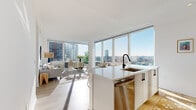 Located at 180 Morgan Street in Jersey City, NJ, this modern one-bedroom, one-bathroom residence offers a thoughtfully designed open-concept layout bathed in abundant natural light. Floor-to-ceiling windows wrap the living and dining areas, framing expansive views of the Jersey City skyline and beyond, creating a bright and airy ambiance throughout.

The kitchen is sleek and functional, featuring white cabinetry with matte black hardware, a waterfall-edge island with an undermount sink, stainless steel appliances, and warm light wood flooring that flows seamlessly into the living and dining areas. A dedicated dining nook beside the oversized windows makes full use of the stunning city views.

The living room is open and inviting, with generous natural light pouring in through the large window panels. The bedroom offers a calm retreat with its own city-facing window, light wood floors, and ample space for a comfortable arrangement.

The bathroom is elegantly finished with white subway tile, a soaking tub with matte black fixtures, a modern vanity with an illuminated mirror, and clean tile flooring — combining style and practicality. Throughout the residence, a consistent palette of white walls, light wood floors, and black accents creates a cohesive, contemporary aesthetic ideal for urban living.