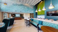 This thoughtfully designed single-floor communal space draws inspiration from coastal living, blending artistic flair with functional amenities throughout its open layout. The most striking feature is a sweeping ocean wave mural adorning the coffered ceiling, rendered in vivid teal and white tones that immediately establish the space's coastal identity. Light-toned herringbone hardwood flooring runs throughout, lending warmth and visual continuity.

A central lounge area anchors the space, featuring a large leather sectional sofa, plush armchairs, and a wall-mounted flat-screen TV backed by dark built-in shelving. Adjacent to this is a communal work and dining counter with white quartz surfaces, tufted aqua banquette seating, leather chairs, and pendant lighting overhead. A wet bar with navy cabinetry, brass hardware, white subway tile backsplash, and a bold living moss wall installation provides a vibrant focal point.

A separate double-height gathering lounge boasts soaring ceilings, floor-to-ceiling windows flooding the space with natural light, a sculptural pendant light fixture, tufted white wall paneling, and curated seating arrangements. Throughout, the cohesive palette of teal, navy, warm leather, and natural greenery creates an inviting, resort-inspired ambiance ideal for socializing or relaxing.