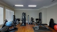 This fitness room offers a dedicated space for strength training and exercise. The room features warm hardwood flooring accented by dark rubber equipment mats, providing both aesthetic appeal and functional protection beneath the machines. Several strength-training stations are arranged throughout the space, including a multi-function cable machine, a leg press or extension unit, and additional resistance machines suited for a variety of muscle groups. The walls are finished in a soft, neutral gray-green tone, complemented by clean white crown molding and baseboards that lend a polished appearance. Two large rectangular fluorescent light fixtures mounted flush to the ceiling ensure the room is well-lit throughout. A window on the left wall allows for natural light to supplement the artificial lighting, contributing to an open and energizing atmosphere. The ceiling height adds to the sense of openness, making the space feel comfortable even with multiple large machines present. Overall, the room is thoughtfully organized to maximize usability while maintaining a tidy, functional environment suitable for regular fitness routines.