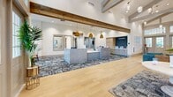 This clubhouse in Orlando, FL, offers a generously sized, single-story community facility designed with a coastal-inspired aesthetic and high-end finishes throughout. The grand entrance welcomes visitors with soaring vaulted ceilings accented by exposed wood beams, light hardwood flooring, and an abundance of natural light streaming through large multi-pane windows and glass-paneled double doors.

The main living lounge is a standout gathering space featuring dramatic cathedral ceilings with a statement chandelier, a wood-plank fireplace wall with a mounted TV, built-in teal cabinetry, and plush sectional seating arranged over a palm-motif area rug. Adjacent to the lounge, an open café-style kitchen bar showcases a teal island with marble countertops, open shelving, and dual mounted screens — ideal for social gatherings.

A dedicated multi-room fitness center is comprehensively outfitted with TRX systems, a punching bag, cable machines, weight benches, treadmills, stair climbers, and cardio equipment, all set on textured carpet with mirrored walls and ample natural light.

Additional areas include a bright multipurpose room with terrazzo flooring and built-in gray cabinetry, a well-appointed mailroom with decorative wallpaper and pendant lighting, and clean, art-lined hallways that connect each space seamlessly throughout the facility.