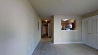 This single-floor residential space offers a clean, functional layout designed for comfortable everyday living. The living area is finished with wall-to-wall carpeting in a neutral tone, creating a warm and cohesive atmosphere throughout. A defining architectural feature is the pass-through kitchen window, which opens directly from the kitchen into the main living area, promoting an open and connected feel between spaces. The kitchen is equipped with stainless steel appliances, including a microwave and range, and is complemented by warm wood-toned cabinetry that adds a welcoming contrast to the light-colored walls. Recessed fluorescent lighting in the kitchen ensures a bright and practical workspace. The neutral beige walls throughout the unit provide a versatile backdrop, allowing for a variety of interior styling choices. A hallway extends from the main living space, leading to additional rooms, including what appears to be a bathroom and at least one other room, offering a sense of privacy and separation from the main living areas. The overall ambiance of the space is understated and move-in ready, with a straightforward design that prioritizes functionality and livability.
