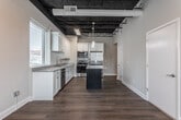 Located on 13th Street in the heart of Columbus, Georgia, this single-floor loft-style unit blends industrial character with modern finishes. A defining architectural feature is the dramatic exposed black ceiling with visible ductwork and steel framing, which contrasts sharply with the clean white walls and warm hardwood-style flooring found throughout the main living areas.

The kitchen is a standout space, outfitted with crisp white shaker cabinetry, granite countertops, a subway tile backsplash, and a full suite of stainless steel appliances including a range, built-in microwave, dishwasher, and refrigerator. A dark-toned center island with pendant lighting above provides additional workspace and a focal point for the room. Large windows allow natural light to pour in, offering views of the surrounding downtown Columbus skyline.

The bedroom features plush carpet flooring, a ceiling fan, and a generous window with city views. The full bathroom is efficiently designed with a white vanity, granite countertop, a glass-enclosed walk-in shower with tile surround, and wood-look flooring. A dedicated laundry closet houses a stacked Whirlpool washer and dryer, along with an electrical panel and storage shelving. The entry features a smart-lock door and clean, neutral finishes throughout.