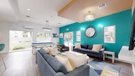 Located at 11615 Firestone Blvd in Norwalk, CA, this single-floor community clubroom is a thoughtfully designed social and recreational space. The interior makes a bold visual statement with a striking teal accent wall, contrasted against crisp white walls and warm wood-toned ceiling panels and wall cladding. Wood-look plank flooring runs throughout, adding warmth and continuity to the space.

The lounge area is generously furnished with multiple navy and gray upholstered sofas and accent chairs, arranged around a round white coffee table atop a geometric, multi-colored area rug. A wall-mounted flat-screen TV is set against a natural wood panel feature wall, creating a comfortable media viewing zone. Decorative sconce lighting, a circular mirror, and curated artwork contribute to the polished, modern aesthetic.

A full-size pool table with a vivid blue felt surface serves as a central recreational feature, positioned near large sliding glass doors and windows that flood the space with natural light and offer views of an outdoor pool area. A wall-mounted cue rack adds a functional, stylish touch nearby.

A glass-enclosed office or workspace is visible from the main lounge, providing a quieter, private setting within the broader space. The combination of recreational amenities, comfortable seating, and contemporary design makes this clubroom a well-rounded community gathering destination.