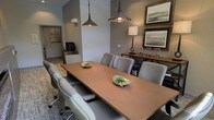 This single-floor space is designed as a functional and refined meeting environment. At its center sits a rectangular wood-toned conference table surrounded by cushioned gray rolling chairs, comfortably accommodating multiple attendees. Two industrial-style pendant lights hang overhead, casting warm, focused illumination across the table, which is accented with small decorative greenery bowls. The walls feature a textured wallcovering in neutral tones, complemented by framed landscape artwork that adds a polished, professional aesthetic. A credenza along one wall holds a table lamp and decorative accessories, while a printer or equipment station is visible through a connecting hallway, indicating practical office functionality nearby. The flooring transitions between carpet in the main meeting area and what appears to be wood-look plank flooring in the adjacent corridor. Wall sconces provide additional ambient lighting throughout. The overall ambiance is modern yet approachable, blending clean lines with warm finishes to create a space that feels both professional and welcoming — well-suited for focused discussions, presentations, or collaborative work sessions.