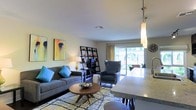 This single-floor residence offers a welcoming and well-appointed open-concept layout that seamlessly connects the living and kitchen areas. The living room is tastefully designed with a comfortable gray sectional sofa accented by vibrant teal throw pillows, a mid-century modern round coffee table, and a stylish geometric-patterned area rug in muted yellows and grays. A charcoal wingback accent chair adds a sophisticated touch, while a sleek black shelving unit serves as both a functional and decorative focal point. Colorful abstract artwork adorns the walls, contributing to the space's contemporary aesthetic. Natural light pours generously into the room through large sliding glass doors that open directly onto an outdoor patio, offering a seamless indoor-outdoor connection with views of lush greenery beyond. The kitchen area features granite or stone countertops and a modern pendant light fixture, hinting at quality finishes throughout. Smooth white ceilings with recessed ventilation maintain a clean, airy feel. Overall, the space balances modern design with a warm, livable ambiance, making it an inviting environment suited for both relaxing and entertaining.