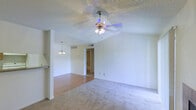 This single-floor space features an open-concept living area that combines two distinct flooring types: smooth hardwood-style planks in the dining or kitchen-adjacent zone and plush wall-to-wall carpeting in the main living area, creating a clear visual separation between functional zones. The textured popcorn ceiling adds a classic touch and houses a multi-blade ceiling fan with an integrated light fixture, providing both ventilation and overhead illumination. A pendant light fixture is suspended nearby, likely marking the transition toward a dining or kitchen area, which is partially visible and appears to include a countertop or bar-style divider. The walls are painted in a clean, neutral white tone, enhancing the sense of brightness throughout. A doorway visible from the living area leads to what appears to be a bathroom or utility space, with tiled flooring and shelving hinting at additional storage. Recessed HVAC vents are positioned along the walls and ceiling, ensuring climate control throughout the space. Multiple electrical outlets are distributed along the baseboards, offering practical functionality. Overall, the space presents a clean, functional layout with a bright, open ambiance.