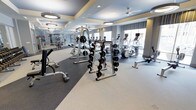 This fitness center offers a spacious, single-level layout designed to accommodate a comprehensive workout experience. The space is thoughtfully divided into distinct zones for cardio and strength training. Along the perimeter, large windows allow generous natural light to fill the room, complemented by modern pendant lighting and ceiling fans that maintain a comfortable atmosphere during workouts.

The strength training area is well-stocked with a barbell rack holding an organized selection of weighted bars, a cable machine, free weights, and a dedicated bench press station. The flooring transitions from dark rubber matting in the weightlifting zone to lighter flooring in the cardio area, providing both safety and visual distinction between workout spaces.

The cardio section features a row of modern elliptical trainers and stair climbers, each equipped with individual display screens. Full-length mirrors line one wall, enhancing the sense of openness and allowing users to monitor their form. The overall ambiance is bright, clean, and professional, making the fitness center a functional and inviting space for a wide range of exercise routines.