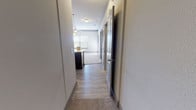 This single-floor unit is thoughtfully laid out, connecting its various living spaces through a central hallway finished with wood-look luxury vinyl plank flooring. The hallway leads from the entry corridor into the main living and kitchen area, with doors branching off to bedrooms and a bathroom along the way.

The kitchen is designed for functionality, featuring dark cabinetry that extends to the ceiling, stainless steel appliances including a full-size refrigerator and range, a stainless steel sink, and light-toned countertops. A peninsula with pendant lighting above opens directly to the living area, creating a seamless flow between cooking and entertaining spaces. The living area is finished with plush carpeting and benefits from natural light provided by windows overlooking outdoor greenery.

The bedrooms are carpeted and fitted with ceiling fans, offering comfortable, well-lit retreats. Crown molding frames the ceilings throughout, adding a refined architectural detail. One bedroom includes a spacious walk-in closet outfitted with built-in shelving and hanging rods for ample storage.

The bathroom is clean and modern, featuring wood-look vinyl plank flooring, a soaking tub with a tiled surround and curved shower rod, a toilet, and a dark-cabineted vanity with a light countertop. The overall palette of neutral tones throughout creates a cohesive, move-in-ready atmosphere.