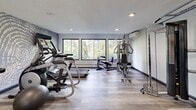 Located at 12550 Portland Ave in Burnsville, Minnesota, this well-appointed fitness center offers a comprehensive exercise environment within a compact, efficiently utilized single-room layout. The space features light wood-style flooring throughout, neutral gray walls, and a textured ceiling with recessed lighting that keeps the room bright and inviting.

A striking decorative wall mural with an abstract wood-grain pattern serves as a focal point, adding a modern aesthetic touch to the interior. Large windows along one wall flood the space with natural light and offer views of the surrounding greenery, creating an energizing atmosphere for workouts.

The fitness center is equipped with a well-rounded selection of equipment, including treadmills, an elliptical trainer, and a stationary bike for cardiovascular exercise. A Matrix dual cable machine provides versatile strength training options, while a multi-station weight machine supports a variety of resistance exercises. A tiered dumbbell rack stocked with a full range of free weights rounds out the strength training offerings. A wall-mounted television is positioned for easy viewing during cardio sessions.

The space also features a wall-mounted air conditioning unit for climate control, wall sconces for ambient lighting, and double glass doors at the entrance. Fitness center rules are posted prominently, ensuring a safe and organized environment for all users.