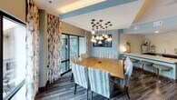 This thoughtfully designed single-floor amenity space offers a sophisticated blend of modern aesthetics and functional comfort, ideal for gathering and entertaining. The open lounge area sets a refined tone with soft blue-grey walls, dark hardwood-style tile flooring, and a striking textured accent wall. Curated furnishings — including a plush sofa, accent chairs, and bold abstract artwork — create an inviting atmosphere bathed in recessed lighting. Adjacent to the lounge, the dining area serves as a centerpiece for social gatherings, anchored by a large rectangular wood table surrounded by woven upholstered chairs. A dramatic cluster chandelier with exposed Edison-style bulbs hangs overhead, while floor-to-ceiling windows dressed in patterned drapes flood the room with natural light and provide views of the surrounding exterior. A wall-mounted flat-screen TV adds a modern, functional touch. The kitchenette is outfitted with dark wood cabinetry, light quartz countertops, a textured tile backsplash, stainless steel appliances including a built-in microwave and warming drawer, and a double-basin sink — offering full convenience for catered events or casual use. Together, these areas form a cohesive, elegantly appointed communal retreat.