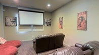 This single-level home theater room is designed to deliver a cinematic experience from the comfort of home. The centerpiece of the space is a large motorized projection screen mounted on the front wall, providing an expansive viewing surface ideal for movies, sports, or gaming. Recessed ceiling lighting offers subtle, controllable illumination that complements the theater ambiance without causing screen glare.

The seating consists of dark brown leather reclining sofas arranged to face the screen directly, providing comfortable, lounge-style viewing for multiple guests. The carpeted flooring adds warmth and acoustic dampening, enhancing the overall sound quality within the room.

The walls are finished in a neutral tan tone, creating a understated backdrop that keeps the focus on the screen. Cinema-themed wall art, including a classic Hollywood clapperboard sign and a vintage movie poster, reinforce the room's dedicated entertainment purpose and add character to the space.

Overall, the room strikes a balance between functional home theater design and inviting comfort, making it a well-appointed space for entertainment gatherings or private movie nights.