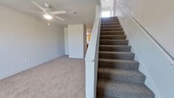 This two-story home offers a practical and well-organized layout across two floors. The main floor comprises the larger share of the living space and includes a kitchen equipped with light wood cabinetry, a gas range, over-the-range microwave, and a full-size refrigerator — all in a coordinated black finish. Durable wood-look flooring runs through the kitchen area, while plush carpet covers the main living space, which features a ceiling fan with light fixture. A carpeted staircase with a white-painted banister connects the main floor to the upper level. The upper floor is more compact and hosts the three bedrooms and two full bathrooms. Each bedroom features soft carpet flooring; the bedrooms include ceiling fans with light fixtures and windows that allow natural light. Several bedrooms are equipped with double-door closets offering generous storage. Both bathrooms are identically appointed with wood-look flooring, warm natural wood vanities with gray countertops, large mirrors, a toilet, and a tiled shower-tub combination. The home maintains a clean, neutral color palette with light gray walls and white trim throughout, creating a bright and cohesive atmosphere across all rooms.