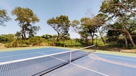 This amenity space combines outdoor recreation with refined interior design to create a welcoming, multi-functional facility. Outside, a full-size tennis court finished in a classic blue surface with crisp white court markings and a regulation net is set against a lush, tree-lined backdrop under open skies, offering a serene and private setting for play.

Inside, the interior spaces are thoughtfully designed with warm hardwood-style flooring and exposed natural wood ceiling beams that lend a rustic yet sophisticated character throughout. The central lounge area is anchored by a dramatic floor-to-ceiling stacked stone water feature, which serves as a stunning focal point. Flanking the feature are cushioned wooden armchairs with navy blue accent pillows, arranged around a round coffee table atop a woven area rug — creating an inviting space for relaxation.

The open common areas flow seamlessly, with recessed lighting illuminating the neutral white walls adorned with coastal-themed artwork. A passenger elevator provides convenient access, while automatic glass-panel doors connect interior zones with ease. Natural light filters in through windows fitted with plantation shutters, enhancing the bright, airy ambiance. High-top seating areas offer casual gathering spots, making this facility ideal for both active recreation and social engagement.