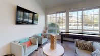 Located at 20155 San Joaquin Terrace in Ashburn, Virginia, this single-floor community amenity space is designed to serve as a social and recreational hub. The open layout is unified by striking herringbone-patterned wood flooring and crisp white walls accented with crown molding, creating a clean, modern aesthetic throughout.

The game lounge area is a standout feature, equipped with a shuffleboard table, a wall-mounted oversized Scrabble board, and a Jenga tower set atop a round coffee table. Comfortable lounge seating with colorful accent pillows is arranged to encourage conversation and casual gatherings, while a wall-mounted flat-screen TV adds to the entertainment options. Large, grid-pane windows flood the space with natural light and offer views of the surrounding outdoor greenery.

The kitchenette is thoughtfully outfitted with white cabinetry, warm beige countertops, stainless steel appliances including a beverage refrigerator and dishwasher, a sink, and a coffee station. A large flat-screen TV is mounted above the counter, and pendant lighting and curated wall art add a polished, welcoming touch.

Together, these areas create a versatile, well-appointed communal environment ideal for relaxed socializing, recreational activities, and everyday gatherings in the heart of Ashburn.