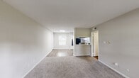 This single-story unit is designed with a functional, open layout that connects the main living spaces seamlessly. The living area is generously sized, finished with neutral-toned carpet and light gray walls that create a clean, understated aesthetic. A front entry door is flanked by a window that draws in natural light, while a second window at the far end of the space adds additional brightness throughout. The layout transitions naturally toward the kitchen, which is accessible via a pass-through opening and a separate entry. The kitchen is well-appointed with white cabinetry, white subway tile backsplash, marble-style countertops, and a full suite of black appliances, including a refrigerator, dishwasher, and electric range. Wood-look flooring grounds the kitchen with a warm contrast to the cabinetry. The bedroom is a well-proportioned, carpeted room with a window offering outdoor views and ample wall space. The bathroom features a dual-sink vanity with marble-style countertops, white cabinetry, and a hallway leading to additional spaces including a closet and shower area. Wood-look flooring extends through the bathroom and hallway, maintaining a cohesive look throughout the unit.