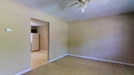 This single-floor residential space offers a straightforward and functional layout designed for comfortable everyday living. The main living area is finished with wall-to-wall carpeting in a neutral beige tone, creating a warm and understated atmosphere. A ceiling fan with a light fixture is centrally mounted on the textured ceiling, providing both airflow and ambient lighting to the room. The walls are painted in a soft, muted tone that complements the light flooring and trim throughout. Clean white baseboards line the perimeter of the room, adding a crisp finishing detail. An open doorway connects the living area directly to the kitchen, which features smooth, easy-to-clean flooring and is equipped with a full-size white refrigerator. A dark appliance, likely a stove or microwave, is also visible in the kitchen area, indicating a practical and equipped cooking space. The transition between the carpeted living room and the kitchen's hard flooring is defined by the open archway, creating a clear yet connected separation between the two functional areas. Overall, the space presents a clean, move-in ready environment with a simple, no-frills design aesthetic suitable for a variety of occupants.