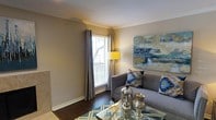 This single-floor space showcases a thoughtfully designed living room that balances comfort and contemporary style. The room features warm, neutral-toned walls complemented by crisp white crown molding, adding a refined architectural detail that draws the eye upward. Dark hardwood flooring grounds the space and contrasts elegantly with the lighter walls and furnishings.

A large window dressed with floor-length grommet-style curtains allows an abundance of natural light to filter in through horizontal blinds, creating a bright and welcoming atmosphere. The living area is anchored by a grey upholstered sofa adorned with decorative Ikat-patterned throw pillows in blue and gold tones. A glass-topped coffee table with a metallic frame sits at the center, styled with silver and bronze decorative vessels.

A striking large-scale abstract canvas painting in shades of blue, teal, and gold serves as a bold focal point on one wall, adding a vibrant artistic element to the space. A gold tripod floor lamp provides warm ambient lighting. A teal geometric-patterned area rug ties the color palette together, contributing to the cohesive and polished overall ambiance.