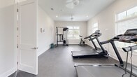 This fitness facility is designed to support a comprehensive workout experience within a single-level layout. The space is finished with durable dark flooring throughout, providing a practical and easy-to-maintain surface well-suited for exercise. Crisp white walls and recessed lighting keep the environment bright and clean, while ceiling fans promote air circulation during workouts.

Multiple windows fitted with white-slatted blinds line the walls, allowing natural light to filter in and offering views of the surrounding outdoor greenery. French doors at one end of the room open to an adjacent outdoor area, enhancing ventilation and providing a seamless connection to the exterior.

The facility is equipped with a variety of cardio machines, including treadmills and a stationary spin bike, alongside a multi-function cable strength machine, an adjustable weight bench, and a dumbbell rack stocked with free weights. The layout is open and spacious, allowing ample room to move freely between equipment stations. An emergency exit door and fire safety features are also present, reflecting attention to safety standards. Overall, the space offers a clean, functional, and well-lit environment suited for diverse fitness routines.