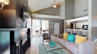 This single-floor space makes a strong first impression with its dramatic vaulted ceilings that soar overhead, creating an expansive, airy atmosphere throughout the main living area. A ceiling fan is centrally positioned to complement the open feel, while natural light pours in through large windows fitted with horizontal blinds, illuminating the light tile flooring below. The living area is anchored by a bold, dark fireplace with a mounted flat-screen television above, serving as a striking focal point against the warm grey walls accented with white crown molding. Seating is arranged thoughtfully around a sleek glass coffee table atop a teal geometric-patterned area rug, with a grey sofa dressed in vibrant yellow and teal accent pillows adding a lively pop of color. A patterned accent chair adds additional character to the space. Just beyond the main living area, a casual dining nook sits beside the windows, featuring bar-height seating and an inviting glimpse of an outdoor area. A vanity area with a mirror is visible nearby, hinting at well-appointed additional spaces. The overall ambiance is modern, cheerful, and thoughtfully designed, balancing comfort with style.