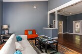 This single-story space in Macon, Georgia, is designed to function as a professional office or community-facing facility, combining a functional reception area with a comfortable lounge. The interior is finished with warm hardwood flooring throughout and painted in a consistent blue-gray tone, creating a cohesive and calming atmosphere across the entire space.

The entry area features a glass-paneled front door flanked by sidelight windows, allowing natural light to filter into the space. A workstation with a desk and seating is positioned near the entrance, serving as an operational hub. Two white service counters with pass-through windows define the reception zone, offering a structured and organized point of contact for visitors.

The lounge area transitions naturally from reception and is distinguished by vaulted ceilings with exposed white beams, adding architectural character and a sense of openness. A set of French doors at the rear provides views and access to an outdoor pool area, further enhancing the brightness of the room. The lounge is furnished with a neutral sofa, vibrant orange accent chairs, a glass-top coffee table displaying reading materials, and a colorful area rug, creating an inviting and lively gathering space.