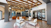 Located at 2870 Pearsons Pky in Plainfield, Indiana, this expansive single-floor community clubhouse offers a generously sized, open-plan amenity space designed for socializing and recreation. The interior is defined by a striking coffered ceiling with rich wood beam detailing and an array of Edison-style pendant lights that lend the space a warm, industrial-modern aesthetic. Large-format tile flooring runs throughout, complementing the neutral gray and white color palette accented by warm wood tones and brick column features.

The main lounge area provides multiple seating arrangements, including cream upholstered armchairs, leather sofas, and a bar-height counter with leather stools positioned beneath pendant lighting and a wall-mounted television — ideal for gathering to watch sports or events. Full-height sliding glass doors along the perimeter flood the interior with natural light and offer direct views of and access to the outdoor courtyard.

A dedicated games area features two billiards tables, a wall-mounted TV, and a separate alcove with an arcade cabinet and a basketball shooting game. A private room with glass-panel doors provides a quieter, flexible space. Throughout, the design balances energetic recreational functionality with comfortable, inviting lounge areas, creating a well-rounded communal environment.