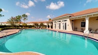 This single-story Mediterranean-inspired facility is defined by its warm stucco exterior, distinctive terracotta barrel-tile roof, and classic architectural detailing that evokes a refined Florida resort aesthetic. The centerpiece of the property is a generously sized, freeform swimming pool with crystal-clear turquoise water, accented by a cobalt blue tile border and surrounded by a spacious brick-paver deck ideal for lounging. Cushioned chaise lounges are positioned poolside, offering a comfortable setting beneath the open sky.

The adjacent clubhouse structure is distinguished by a series of large French doors that line the façade, allowing abundant natural light to flow between the interior and the outdoor pool area. Wall-mounted lantern-style light fixtures flank the entry, adding a refined decorative touch. The building's clean, neutral stucco finish complements the overall Mediterranean character of the complex.

Mature pine trees and lush tropical landscaping frame the perimeter, providing a natural backdrop that enhances the sense of a private, resort-like retreat. The surrounding residential buildings share the same architectural language, contributing to a cohesive and well-appointed community environment. The entire outdoor area benefits from ample Florida sunshine, creating a bright and inviting atmosphere throughout the day.