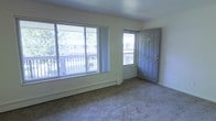 This single-floor residential space offers a clean, functional layout centered around a bright and open living area. The room is finished with wall-to-wall carpeting that adds a sense of warmth underfoot, while light-toned walls create a neutral and inviting atmosphere throughout.

Natural light is a defining feature of the living area, which is equipped with a large sliding glass window and a separate entry door — both overlooking an outdoor balcony. The balcony provides pleasant views of surrounding trees and neighboring structures, bringing a touch of greenery and openness to the interior. The generous window arrangement ensures the space remains well-lit throughout the day.

A baseboard heating unit runs along the exterior wall beneath the windows, offering an efficient and unobtrusive heating solution. The entry door is solid and paneled, providing both security and a classic architectural detail. The overall design of the space is straightforward and practical, with clean lines and minimal ornamentation that make it easy to personalize and furnish. The single-floor layout ensures seamless accessibility and flow, making it a comfortable and livable environment.