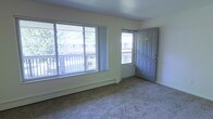 This single-floor residential space offers a clean, functional layout centered around a bright and open living area. The room is finished with wall-to-wall carpeting that adds a sense of warmth underfoot, while light-toned walls create a neutral and inviting atmosphere throughout.

Natural light is a defining feature of the living area, which is equipped with a large sliding glass window and a separate entry door — both overlooking an outdoor balcony. The balcony provides pleasant views of surrounding trees and neighboring structures, bringing a touch of greenery and openness to the interior. The generous window arrangement ensures the space remains well-lit throughout the day.

A baseboard heating unit runs along the exterior wall beneath the windows, offering an efficient and unobtrusive heating solution. The entry door is solid and paneled, providing both security and a classic architectural detail. The overall design of the space is straightforward and practical, with clean lines and minimal ornamentation that make it easy to personalize and furnish. The single-floor layout ensures seamless accessibility and flow, making it a comfortable and livable environment.