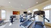 Located at Ashton Village in Westerville, Ohio, this fitness center offers a well-equipped and inviting space designed to support a variety of workout routines. The single-floor facility features a clean, neutral color palette with warm beige walls, crown molding, and recessed lighting that creates a bright and comfortable environment for exercise.

The cardio area is outfitted with multiple treadmills and an elliptical trainer from Life Fitness, providing residents with quality equipment for cardiovascular workouts. A wall-mounted flat-screen television is positioned for easy viewing during exercise sessions. A stationary bike adds further variety to the cardio options available.

The strength training area includes a dedicated free weight station with a tiered dumbbell rack and an adjustable workout bench, accommodating a range of resistance training exercises. The carpeted flooring throughout the space adds comfort underfoot and contributes to a quieter workout atmosphere.

Natural light flows in through elegant arched windows fitted with plantation shutters, giving the space an airy, upscale feel. A large decorative artwork piece on the wall adds a touch of style to the functional environment. Overall, the fitness center strikes a balance between practicality and polished design, making it an appealing amenity for residents of Ashton Village.