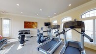 Located at Ashton Village in Westerville, Ohio, this fitness center offers a well-equipped and inviting space designed to support a variety of workout routines. The single-floor facility features a clean, neutral color palette with warm beige walls, crown molding, and recessed lighting that creates a bright and comfortable environment for exercise.

The cardio area is outfitted with multiple treadmills and an elliptical trainer from Life Fitness, providing residents with quality equipment for cardiovascular workouts. A wall-mounted flat-screen television is positioned for easy viewing during exercise sessions. A stationary bike adds further variety to the cardio options available.

The strength training area includes a dedicated free weight station with a tiered dumbbell rack and an adjustable workout bench, accommodating a range of resistance training exercises. The carpeted flooring throughout the space adds comfort underfoot and contributes to a quieter workout atmosphere.

Natural light flows in through elegant arched windows fitted with plantation shutters, giving the space an airy, upscale feel. A large decorative artwork piece on the wall adds a touch of style to the functional environment. Overall, the fitness center strikes a balance between practicality and polished design, making it an appealing amenity for residents of Ashton Village.