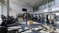 This state-of-the-art fitness center offers a spacious, open-concept workout environment designed to accommodate a wide range of training styles and fitness goals. The facility features soaring double-height ceilings with a large industrial ceiling fan, creating excellent airflow and an airy atmosphere throughout the space. Floor-to-ceiling glass windows along one side of the room flood the interior with natural light, enhancing the bright and energizing ambiance. The polished concrete flooring is both durable and visually sleek, complementing the modern grey-and-white aesthetic of the walls and ceiling. The strength training area is outfitted with professional-grade equipment, including a multi-functional power rack, cable machines, free weights, and a Rogue plyo box, catering to functional and strength-based workouts. A dedicated cardio section lines the perimeter with machines including rowing machines and incline trainers. Wall-mounted mirrors expand the sense of space and allow users to monitor their form. A wall-mounted flat-screen television adds an entertainment element to the workout experience. The overall design strikes a balance between high performance and contemporary style, making it a well-rounded facility suited for serious athletes and casual fitness enthusiasts alike.