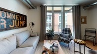 This well-appointed single-floor space showcases a thoughtfully designed open-concept layout that blends functionality with contemporary style. The kitchen serves as a central highlight, featuring stainless steel appliances including a refrigerator, range, and microwave, complemented by warm wood cabinetry and sleek dark countertops. A built-in open shelving unit adds both storage and visual interest, while a peninsula with a breakfast bar and room for multiple stools creates an inviting space for casual dining or entertaining. Industrial-style pendant lights hang overhead, adding a modern, urban character to the kitchen area. Exposed ductwork along the ceiling reinforces the loft-inspired aesthetic throughout the space. The living area is bright and comfortable, benefiting from large floor-to-ceiling windows that invite natural light and offer views of the surrounding neighborhood. A private balcony is accessible directly from the living room, extending the living space outdoors. Hardwood-style flooring flows consistently throughout, tying the rooms together cohesively. The bathroom is clean and well-finished, featuring a wood-toned vanity with dark stone countertops, a large mirror, and a combination tub and shower. The overall ambiance is modern and urban, with an industrial edge balanced by warm wood tones and neutral finishes.