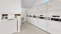 Located at 655 Baker St in Costa Mesa, California, this community laundry facility is designed with both functionality and comfort in mind. The single-floor space features a clean, modern aesthetic defined by crisp white walls, vaulted ceilings, and polished terrazzo-style flooring that reflects natural light throughout the room. A row of top-load washing machines lines one wall, built into sleek white cabinetry with wood-accented countertops, while a bank of stacked dryers occupies an adjacent wall, providing ample capacity for a large residential community. A dedicated folding counter with a dark wood surface runs along the front of the space near a large window, offering views of the landscaped outdoor courtyard and allowing natural light to fill the room. The entrance area features a glass door that opens directly to an outdoor walkway flanked by stone-accented columns, adding a charming architectural touch. Recessed lighting illuminates the interior evenly, complementing the bright, airy atmosphere. The space is well-organized and efficiently laid out, ensuring ease of use. Overall, the facility strikes a balance between practicality and a welcoming, well-maintained environment suitable for daily use.