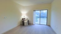 This single-floor space features a thoughtfully designed living area finished with soft, wall-to-wall carpeting that adds warmth and comfort underfoot. The room is characterized by its clean, neutral-toned walls in a soft cream hue, lending a calm and understated aesthetic throughout. A prominent sliding glass door serves as a key architectural feature, drawing in abundant natural light and providing direct access to a private balcony enclosed by a metal railing. The outdoor view reveals lush greenery, offering a pleasant and serene backdrop visible from within the living space. The ceiling incorporates a visible ventilation register, indicating a central air system, while subtle recessed trim work frames the upper walls, adding a refined architectural detail. The overall ambiance is open, airy, and inviting, with neutral finishes that provide a versatile backdrop suitable for a variety of furnishing styles. The combination of natural light from the expansive glass door and the soft carpeted flooring creates a comfortable, livable environment that feels both functional and welcoming.