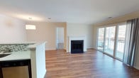 This single-floor space offers an open, flowing layout centered around a well-connected living area. The flooring throughout is a warm-toned hardwood-style laminate that runs continuously across the main living spaces, lending a cohesive and clean aesthetic. Crown molding lines the ceilings, adding a polished architectural detail to the otherwise understated design.

The living area serves as the focal point of the space, anchored by a classic fireplace with a white mantel and black surround. Natural light fills the room through a sliding glass door that opens to a private balcony, offering views of the surrounding outdoor environment. This connection to the exterior enhances the sense of openness throughout.

Adjacent to the living area, a half-wall peninsula defines the kitchen zone, which includes dark cabinetry and a stainless steel refrigerator. The layout allows for easy interaction between the kitchen and living spaces. A dedicated entry hallway leads to the front door, with a utility access panel integrated into the wall. Additional rooms branch off from the main living area, accessible through standard six-panel white doors. The overall ambiance is bright, neutral, and move-in ready.