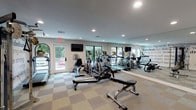 This fitness center is a comprehensively outfitted facility designed to support a wide range of workout routines. The main exercise area is anchored by a lineup of Matrix-brand cardio equipment, including treadmills, ellipticals, and a stationary bike, all arranged in front of a full-length mirror wall that enhances the sense of spaciousness. A multi-use cable strength machine, a flat workout bench, and a rack of dumbbells round out the strength training offerings. A dedicated spin studio provides a focused space for cycling workouts, equipped with multiple indoor cycling bikes, medicine balls, and stability balls, along with a wall-mounted flat-screen TV and large windows that invite natural light and outdoor views. Throughout the facility, recessed lighting illuminates the space evenly, complementing the neutral, checkered carpet tile flooring. A standout feature is the motivational typographic wall mural featuring words like "inspire," "commitment," "strength," and "dedicated," adding energy and character to the environment. A water fountain station and open shelving offer practical amenities. An additional open area with clean white walls and accent artwork provides flexible space for stretching or floor exercises, completing this well-rounded fitness destination.