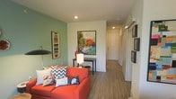 This single-floor space makes a bold visual impression through its thoughtfully curated design and lively color palette. The living area serves as the central gathering point, anchored by a striking coral-red sofa adorned with an eclectic mix of patterned and graphic throw pillows. A sage green accent wall adds warmth and depth, complementing the neutral white walls throughout the rest of the space. A large, vivid abstract painting serves as a prominent focal point, injecting energy and artistic character into the room. A sleek console table with a decorative lamp and small accent pieces provides functional style beneath the artwork. A modern arc floor lamp adds additional ambient lighting to the living area. The flooring consists of wood-style planks that run continuously from the living space into a hallway, creating a cohesive and unified flow throughout the unit. The hallway leads toward what appears to be additional rooms, including a door at the far end suggesting a bedroom or entry. Recessed lighting in the ceiling provides clean, even illumination throughout. A gallery-style wall display near the hallway adds further visual interest. Overall, the space balances contemporary design with vibrant, personality-driven décor.