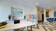 This lobby serves as an elegant and functional welcome space, thoughtfully designed to greet visitors with a polished, coastal-inspired aesthetic. At the center of the room sits a striking marble-topped table with gold-toned legs, providing a refined surface for consultations or informational displays. The floor features a bold geometric herringbone pattern that transitions seamlessly into carpeted areas, adding visual interest and defining the space.

A shiplap accent wall frames the reception counter, which is finished in crisp white cabinetry with a stone countertop. Above the counter, a triptych of abstract coastal artwork in soft teals and aquas introduces a calm, sophisticated color palette that carries throughout the lobby. Recessed lighting illuminates the space evenly, complementing the bright white ceiling and walls.

Display easels positioned throughout the lobby present floor plans, informational brochures, and community imagery, making the space as informative as it is visually appealing. A hallway extends from the main lobby area, offering access to additional areas beyond. The overall ambiance is clean, modern, and approachable — striking a balance between professional functionality and warm, welcoming design.