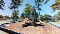This versatile community space combines an inviting outdoor playground with a charming interior clubhouse-style area. Outside, a well-equipped playground structure finished in green and tan stands prominently amid a canopy of mature trees. The multi-level play structure features dual slides, climbing elements, and a covered platform, all set over a wood chip safety surface. A tennis or sport court is visible in the background, adding to the recreational offerings of the outdoor environment.

The interior reflects a warm, cottage-inspired aesthetic, characterized by vertical shiplap-style wall paneling painted in soft white and cream tones. Light hardwood flooring runs throughout, complementing the beamed ceiling detail that adds architectural interest overhead. A comfortable lounge area is anchored by a classic fireplace with a white mantle, creating a cozy focal point. French doors and large windows invite natural light and offer views of the surrounding greenery.

A casual dining area with a round wood table and ladder-back chairs sits adjacent to a wrought iron staircase railing, adding a refined yet relaxed touch. A prominent entry door opens directly to the outdoors, reinforcing the seamless connection between the interior and the lush exterior grounds. Overall, the space presents a functional and welcoming environment suited for community gatherings and recreation.
