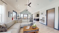 This single-story community space is defined by its striking vaulted ceiling, which rises to a prominent peak accentuated by a sage-green gable wall and dual ceiling fans. Recessed lighting complements the abundant natural light streaming through multiple large windows and glass-paned doors, creating a bright, welcoming atmosphere throughout.

The open-concept layout is organized into several distinct functional zones. A generously sized lounge area anchors the main space, furnished with a large sectional sofa, accent chairs, and a wood-and-metal coffee table atop light-toned luxury vinyl plank flooring. Adjacent to the lounge, a casual dining and gathering zone features round pub-height tables paired with adjustable industrial-style stools, ideal for social interaction.

A fully equipped kitchenette lines one wall, featuring dark cabinetry with gold hardware, white subway tile backsplash, open shelving, a stainless steel refrigerator, microwave, and generous counter space. A glass-partitioned alcove provides a quieter seating nook, while a water fountain and accessible restrooms are conveniently located nearby. Glass doors open directly onto an outdoor patio, seamlessly connecting interior and exterior spaces.