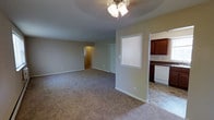 Located at 17 W Chester Pike in Ridley Park, Pennsylvania, this single-floor space offers a straightforward and functional layout well-suited for everyday living. The main living area is generously proportioned, featuring wall-to-wall carpeting in a neutral tone that lends warmth to the room. A ceiling fan with a light fixture overhead provides both comfort and ambient lighting. Soft, neutral-toned walls throughout create a clean and versatile backdrop.

A pass-through window connects the living area to the adjacent kitchen, allowing for an open sightline between the two spaces. The kitchen features rich, dark wood cabinetry, dark countertops, tile flooring, and a white dishwasher, offering a practical cooking and prep environment. The tile flooring in the kitchen contrasts neatly with the carpeted living space.

Multiple windows allow natural light to filter into the unit, brightening the interior. A hallway visible from the main living area leads to additional rooms, suggesting a well-organized floor plan. The overall ambiance is clean, neutral, and move-in ready, making this a practical and comfortable space in the Ridley Park community.