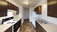 Located at 69 Jadwin Avenue in Richland, Washington, this single-floor unit offers a straightforward, functional layout suited for comfortable everyday living. The floor plan, visible in the dollhouse view, reveals a well-organized arrangement of living spaces, bedrooms, kitchen, and bathroom all on one level.

The living areas are finished with wall-to-wall carpet in a neutral tan tone, complemented by clean white walls and textured popcorn ceilings with centrally placed ventilation. Multiple rooms feature sliding windows that bring in natural light and offer views of the surrounding neighborhood and outdoor scenery.

The galley-style kitchen is equipped with dark wood cabinetry lining both walls, a coil-burner electric range, a full-size white refrigerator, and a double-basin stainless steel sink with a light countertop surface. The kitchen floor features a durable stone-patterned vinyl tile that extends into the bathroom.

The bathroom is well-appointed with a single-basin vanity set into a light countertop, dark wood cabinetry below, a large mirror with overhead lighting, and a full-size tub and shower combination with a curtain rod. The stone-patterned tile flooring ties the wet areas together cohesively.

The bedrooms are generously sized, carpeted, and benefit from natural light through their windows, providing a calm and bright atmosphere throughout this practical Richland residence.