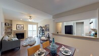 This single-floor residence offers an open-concept design that seamlessly connects the dining area, living room, and kitchen into one cohesive living space. The dining area anchors the layout with a rich, dark wood table surrounded by warm amber chairs, creating an inviting setting for meals and gatherings. A decorative blue glass vase serves as a striking centerpiece, adding a touch of artistic flair.

The living room beyond features a ceiling fan with integrated lighting, a cozy fireplace, and access to an outdoor balcony through sliding glass doors that invite abundant natural light into the space. The open layout is accented by warm neutral tones on the walls and durable hardwood-style flooring throughout, lending the space a modern yet comfortable ambiance.

A pass-through counter connects the living and dining areas to the kitchen, which is equipped with a full-size refrigerator and ample cabinetry. The kitchen's galley-style configuration maximizes efficiency while maintaining visual openness to the rest of the home. Decorative shelving with curated accessories adds personality and depth to the overall interior. The thoughtful layout and cohesive design make this a highly functional and welcoming single-floor living space.