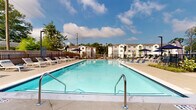 Located at 7326 Wallace Rd in Charlotte, NC, this outdoor amenity space centers around a generously sized swimming pool designed for both relaxation and recreation. The pool features clean, crystal-clear water with easy-access stainless steel entry rails and is framed by a broad concrete sundeck that accommodates an extensive arrangement of white lounge chairs and navy blue market umbrellas, providing ample space for sunbathing and outdoor leisure.

The pool area is enclosed by a combination of wooden privacy fencing and black metal railings, creating a secure and intimate setting. Mature trees surrounding the perimeter contribute natural shade and a lush, green backdrop visible against open skies. A clubhouse structure with a distinctive stone exterior, pitched roofline with exposed timber detailing, and large windows borders one side of the pool, offering an architectural focal point that adds warmth and character to the space.

Multi-story residential buildings are visible in the background, establishing the pool as a central communal feature of the broader community. The overall ambiance is bright and welcoming, with an open-air design that maximizes natural light and outdoor views. The well-maintained landscaping, thoughtful layout, and quality furnishings collectively create a polished, resort-inspired environment well-suited for community gatherings and everyday outdoor enjoyment.