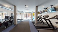 Located at 2500 Summer Lee Dr in Rockwall, Texas, this well-appointed fitness center offers a comprehensive single-floor exercise facility designed to support a wide range of workout routines. The space is bright and open, benefiting from large floor-to-ceiling sliding glass doors that flood the interior with natural light and provide views of the outdoor amenity area beyond.

The fitness center is organized into distinct functional zones. The cardio section features multiple treadmills and rowing machines, accompanied by a row of wall-mounted flat-screen televisions for entertainment during workouts. A dedicated cycling area houses several stationary spin bikes positioned in front of large mirrors, allowing users to monitor their form. A free weight station includes a full rack of dumbbells, while additional strength and resistance machines round out the equipment offerings.

Structural columns with a light wood finish punctuate the space, adding a subtle architectural detail against the clean white walls. Exposed ductwork along portions of the ceiling contributes an industrial design element, while recessed and pendant lighting ensure the space is well-lit throughout. The flooring consists of durable rubberized tiles in contrasting tones, providing safety and comfort underfoot. Double glass doors offer a secondary entrance, and stability balls and resistance bands are also available for functional training.
