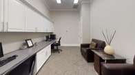This single-floor office space is thoughtfully designed to balance productivity and comfort within a compact, well-organized layout. The room is finished with neutral warm-gray walls, crisp white crown molding, and textured neutral carpeting, creating a professional yet welcoming atmosphere.

A standout feature is the built-in workstation along one wall, comprising an extended gray-topped desk surface paired with an extensive run of white raised-panel cabinetry above. The upper cabinets provide generous enclosed storage, while lower drawers beneath the desk surface offer additional organization. The workstation comfortably accommodates a dual-monitor computer setup along with peripheral equipment, and an ergonomic black task chair completes the workspace.

On the opposite side of the room, a dark brown leather loveseat and accent chair provide a relaxed seating area, each accented with decorative patterned throw pillows. A sleek glass-topped side table adds a refined touch to this lounge corner.

Recessed fluorescent ceiling panels provide consistent, even illumination throughout the space. A paneled white door at the rear leads to an adjacent area, and a wall-mounted thermostat allows for individual climate control. The overall ambiance is clean, functional, and polished.