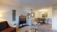 This single-floor residence offers a well-organized layout with distinct living, dining, kitchen, sleeping, and utility spaces. The living room is generously proportioned, featuring plush wall-to-wall carpeting, neutral-toned walls, and a textured ceiling. Natural light filters in through horizontal blinds, creating a bright and welcoming atmosphere. The space flows naturally into a dedicated dining area, which is distinguished by wood-look flooring, a classic chandelier, wainscoting along the lower walls, and room for a full dining set.

The galley-style kitchen is efficiently designed with ample white cabinetry, granite-look countertops, a double basin sink, and stainless steel appliances including a refrigerator and range. Wood-look flooring ties the kitchen neatly together. Adjacent to the kitchen, a dedicated laundry area provides a full-size washer and dryer alongside shelving for storage convenience.

The bedroom offers a comfortable retreat with soft carpeting, windows dressed with curtains for privacy, and sufficient space for a full furniture arrangement. The bathroom features a walk-in shower with tile surround, wood-look flooring, and clean, neutral finishes throughout. Overall, the residence presents a cohesive and functional living environment.