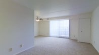 This single-story space features a spacious, open living area designed with simplicity and functionality in mind. The room is finished with wall-to-wall plush carpeting in a neutral beige tone, providing a soft and comfortable underfoot feel while contributing to the overall clean, understated aesthetic.

A prominent sliding glass door dominates one wall, dressed with full-length vertical blinds that allow flexible light control. This feature serves as the primary source of natural light, brightening the interior and offering a visual connection to the outdoors. When the blinds are drawn open, the room is bathed in daylight, enhancing the sense of openness.

The ceiling is smooth and finished in white, maintaining a bright, airy atmosphere throughout. A ceiling fan with an integrated light fixture is centrally positioned overhead, providing both ventilation and ambient lighting — a practical and permanent fixture suited to year-round use.

The walls are painted in a soft, neutral tone that complements the carpeting, creating a cohesive and versatile interior palette. White baseboards frame the perimeter of the room cleanly. The overall layout is open and unobstructed, allowing flexible furniture arrangement and making the space feel generously proportioned.